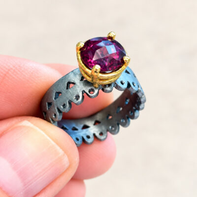 Ring with an 8 mm rhodolite in an 18K gold setting and a rhodium-plated silver openwork band, held between fingers.