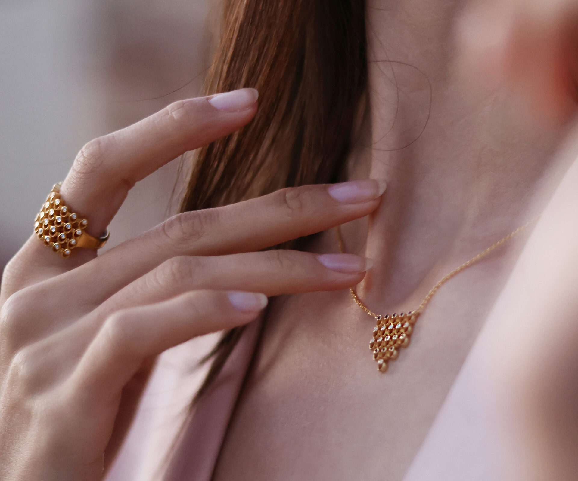 Wide 18K gold diamond ring with a repeating geometric pattern, worn with a matching gold necklace and pale pink jacket.